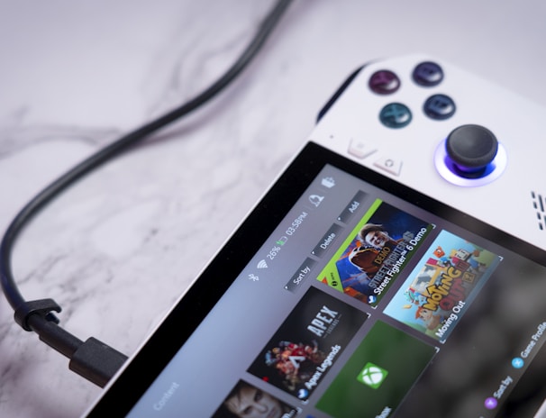 A handheld gaming console with a white body and colorful buttons features a screen displaying various game icons. The device is connected to a power source via a black cable and rests on a marble surface. The interface shows games like Apex and Street Fighter.